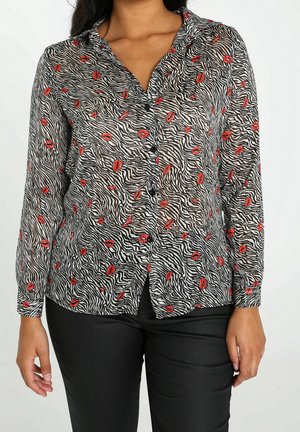 Kvinde iført en skjorte med knapper, der har sorte og hvide zebrastriber samt et mønster med røde læber, kombineret med sorte bukser.