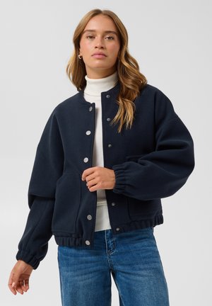Femme portant une veste bleue marine boutonnée sur un pull à col roulé blanc et un jean bleu, debout devant un fond gris clair uni.