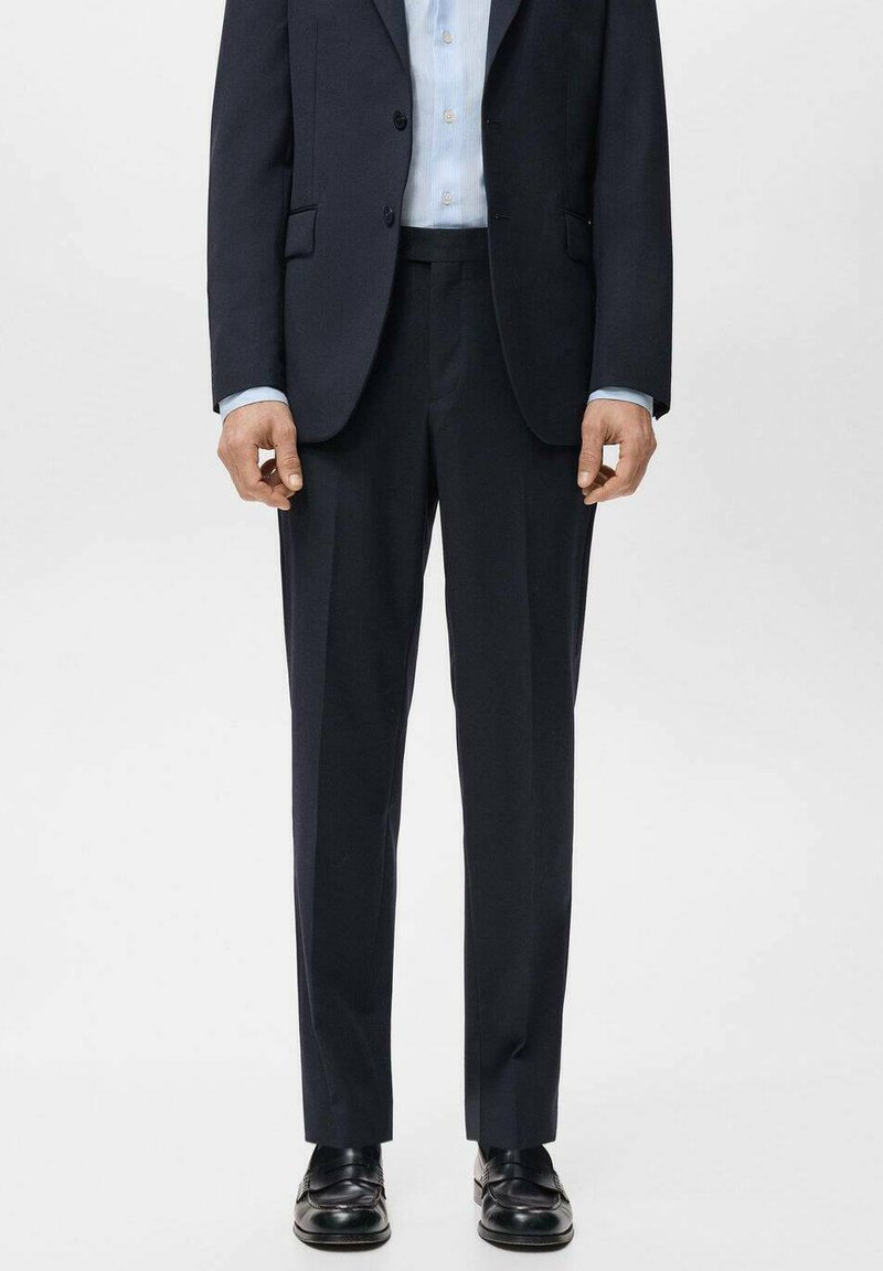 Granatowy garnitur o kroju dopasowanym, z dwurzędową marynarką na dwa guziki, prostymi spodniami oraz błyszczącymi czarnymi butami. Gładka faktura tkaniny.
