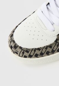 Baskets blanches avec des côtés en toile texturée à motifs noirs et beiges, présentant des perforations et des lacets blancs. Bout arrondi et semelle en caoutchouc.