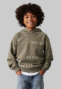 Felpa verde oliva con loghi Calvin Klein di diverse dimensioni, polsini a costine e cappuccio regolabile, abbinata a jeans in denim blu.