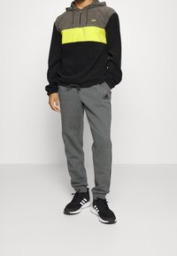 Mand iført en farvet hoodie med gul stribe, grå Adidas sweatpants og sorte Adidas sneakers, stående foran en hvid baggrund.