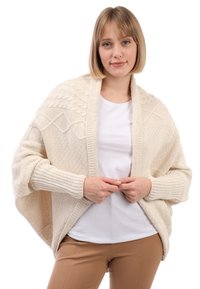 Beżowy sweter dziergany z teksturowanymi wzorami, rozpinany z przodu, z ściągaczami na rękawach, noszony na białej koszuli i zestawiony z jasnobrązowymi spodniami.