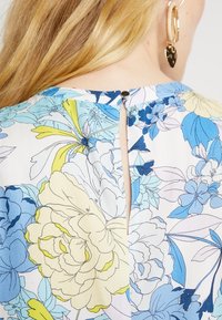 Blouse florale avec des fleurs bleues et jaunes sur un fond clair, dotée d'une ouverture en forme de clé à l'arrière maintenue par un bouton.