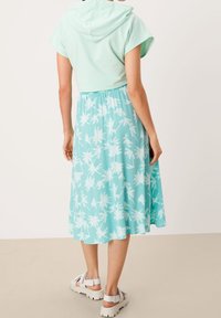 Sweat à capuche vert menthe à manches courtes, associé à une jupe bleu clair ornée d'un motif de feuilles de palmier blanches, porté avec des sandales blanches épaisses.