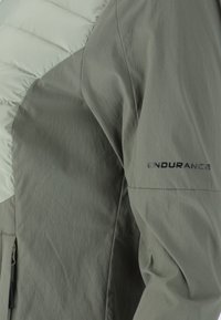 Ljusgrå jacka med en vadderad, isolerad övre sektion och slät nedre tyg; "ENDURANCE"-logotyp synlig på ärmen.