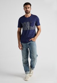 Granatowa bawełniana koszulka z teksturowanym wzorem na klatce piersiowej oraz kieszonką na piersi. Noszona z jasnoniebieskimi dżinsami i białymi trampkami.