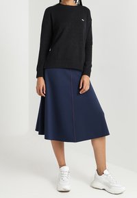 Pull noir en maille avec encolure ronde, associé à une jupe trapèze bleu marine avec une couture rose. Porté avec des chaussures de sport blanches.