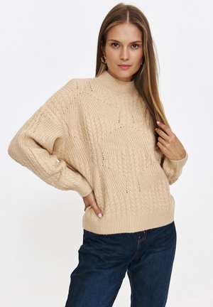 Beżowy sweter dzianinowy z wysokim kołnierzem, wzorem w warkocze, luźnymi rękawami i ściągaczami na końcach rękawów. W połączeniu z ciemnoniebieskimi dżinsami.