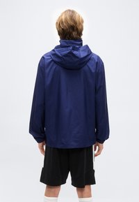 Άτομο που φοράει μπλε μπλε μπλε μπουφάν με κουκούλα, μαύρο σορτς με λευκές ρίγες και λευκές κάλτσες, όρθιο στραμμένο προς τα πίσω σε απλό φόντο.