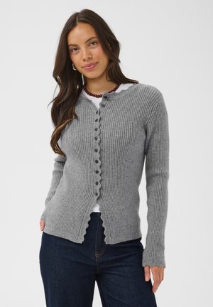Kvinde med langt brunt hår bærer en ribstrikket grå cardigan med knapper over en hvid top, mørke jeans og et perlehalskæde, stående mod en ensfarvet baggrund.