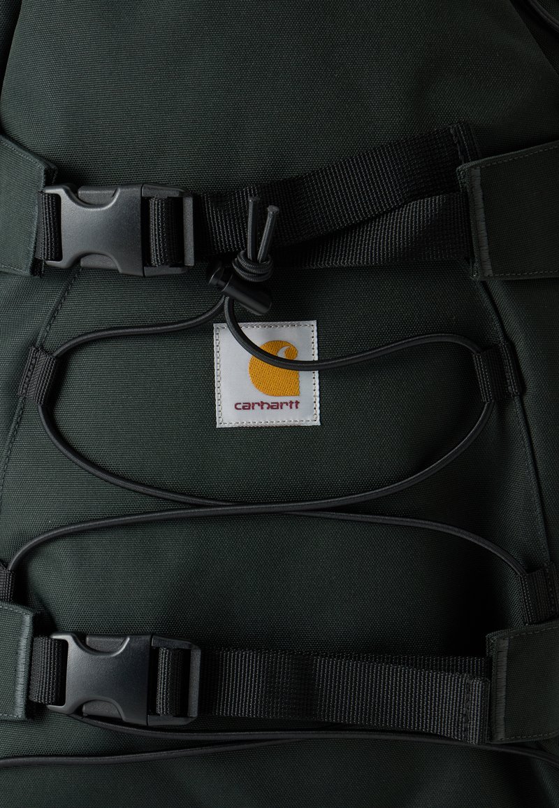 Dunkelgrüner Carhartt-Rucksack in Nahaufnahme zeigt schwarze verstellbare Gurte, Kunststoffschnallen und ein gelbes Carhartt-Logo-Patch.