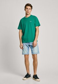 Green cotton t-shirt with short sleeves and crew neck, paired with light blue denim shorts, and black sneakers with cream soles.