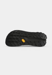 Semelle de chaussure noire robuste avec des rainures profondes et un petit logo jaune au centre, conçue pour l'adhérence et la durabilité.