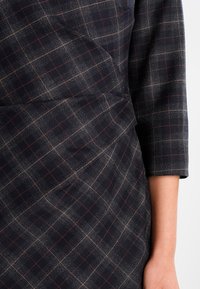 Robe bleu marine avec un motif à carreaux dans des tons de gris et de rouge, présentant des détails drapés et des manches trois-quarts.