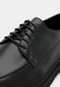 Chaussure en cuir noir à finition lisse, dotée de six œillets ronds et de lacets noirs. Le design présente un profil bas et une silhouette minimaliste.