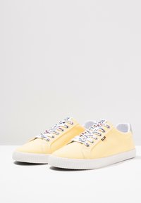 Yellow canvas sneakers with white rubber soles, featuring colorful laces and a branded tongue label. Minimalist design with smooth texture.