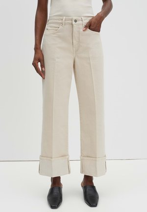 Persoon die beige broek met wijde pijpen en grote manchet draagt, en zwarte slip-on schoenen met puntige neus, staand met één hand in de zak.