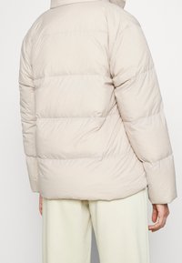 Beige pufferjacka med en quiltad design, med hög hals, långa ärmar och en avslappnad passform, tillverkad av ett mjukt, vattenavvisande material.