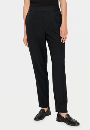 Vrouw die op maat gemaakte, zwarte broek tot aan de enkels en zwarte loafers draagt, staand met de handen ontspannen langs het lichaam.