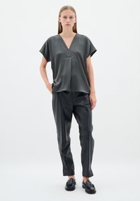 Top gri cu mânecă scurtă, decolteu în V și material texturat, asociat cu pantaloni de piele negri. Modelul stă în fața unui fundal alb simplu.