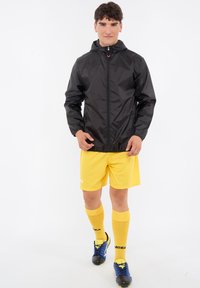 Zwarte windbreaker met rits en capuchon, gedragen over een geel sportuniform met knielange shorts en sokken, met blauwe voetbalschoenen.