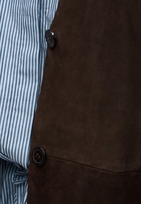 Καφέ ύφασμα από σουέντ με ομαλή υφή, που διαθέτει μαύρα κουμπιά και μερικώς ορατό μπλε και λευκό ριγέ πουκάμισο από κάτω.