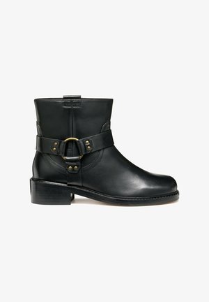 Bota de pele preta até ao tornozelo com bico redondo, tirante lateral com uma fivela circular dourada e um salto baixo em camadas. Textura suave e detalhes mínimos.