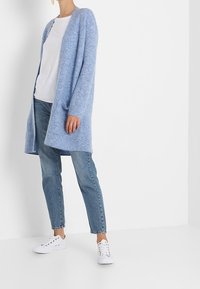 Osoba w jasnoniebieskim długim kardiganie, białej koszulce, niebieskich dżinsach i białych sneakersach, stojąca na tle jednolitego białego tła.