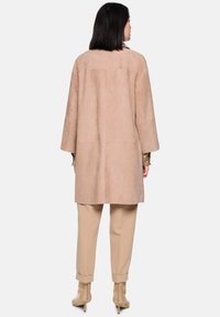 Beiger Wildleder-Langmantel mit lockerem Schnitt, weiten Ärmeln und nahtlosem Design, kombiniert mit beigen, zulaufenden Hosen und hochhackigen Stiefeln.