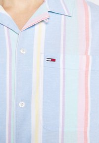 Gros plan d'une chemise à rayures pastel avec boutonnière sur le devant, col, poche poitrine et petit logo rectangulaire rouge, blanc et noir sur la poche.