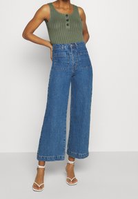 Canotta verde a coste con tre bottoni neri, abbinata a jeans in denim blu a vita alta e ampia, e sandali bianchi con cinturini.