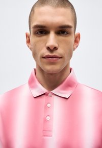 Νεαρός άνδρας με κοντά μαλλιά που φοράει ένα πουκάμισο πόλο σε ροζ και λευκή διαβάθμιση με κουμπιά, κοιτάζοντας μπροστά μπροστά σε απλό φόντο.