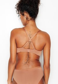 Reggiseno con tessuto nude morbido, design con cinturini, schiena incrociata regolabile e chiusura a gancio sottile. Texture liscia e forma aderente.