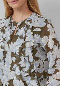 Blumenbluse in Olivgrün mit hellblauem botanischem Druck. Mit Rüschen entlang des Ausschnitts und der Ärmel sowie einem kleinen Schlüsselaugen-Detail.
