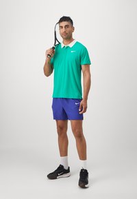 Roheline polo-särk valge kraega, lillad lühikesed püksid, mustad spordijalatsid, hoides tennisereketit, seistes ühtlases taustas.