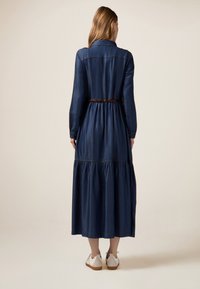 Abito lungo in denim con vita attillata, gonna a strati e design con bottoni. Indossato con una cintura marrone e sneaker bianche.