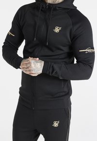 Sudadera negra con cremallera hecha de tela suave, con logotipos dorados, cordón y bolsillos laterales. Combinada con pantalones negros a juego.