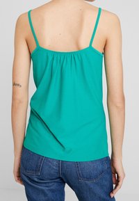 Top sin mangas color turquesa con tirantes finos, con la parte trasera fruncida, hecho de tela suave; combinado con jeans de mezclilla azules.