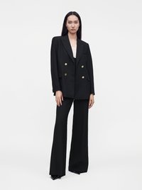 Femme aux longs cheveux foncés portant un blazer noir à double boutonnage avec des boutons dorés et un pantalon large assorti sur fond blanc.