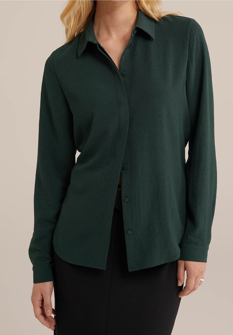 Ciemnozielona koszula z długim rękawem, zapinana na guziki z kołnierzykiem, wykonana z miękkiego, strukturalnego materiału. Posiada luźny krój i zaokrąglony dół.