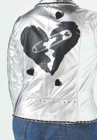 Silberne Kunstlederjacke mit einem großen schwarzen Herzmotiv, Sicherheitsnadeln, kleinen schwarzen Herzapplikationen und metallischen Nieten entlang des Saums.