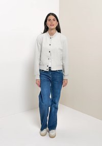 Donna in piedi contro un muro bicolore beige e bianco, indossa una giacca grigio chiaro con bottoni, jeans blu e sneakers bianche.