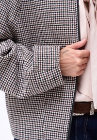 Kurtka w wzór houndstooth w brązowo-białym materiale, charakteryzująca się luźnym krojem, szerokimi mankietami oraz zapięciem na zamek błyskawiczny z przodu.