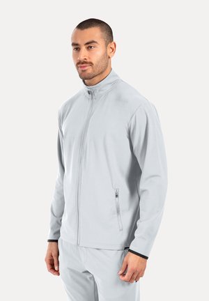 Hombre de pie con chaqueta gris claro con cremallera y mangas largas y pantalones a juego gris claro sobre un fondo liso.