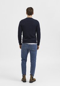 Marineblauer Strickpullover mit Kabelmuster, Rundhalskragen, gerippten Ärmeln, kombiniert mit blauen Jeans und braunen Stiefeletten, von hinten betrachtet.