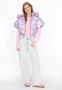 Lawendowa kurtka puchowa z błyszczącego materiału, krótki fason i futrzane akcenty na kapturze, w połączeniu z różową bluzką z długim rękawem oraz jasnymi dżinsami.
