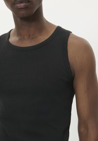Primo piano di una persona che indossa una canotta nera a coste, che mostra parte del petto, della spalla e del collo contro uno sfondo semplice.