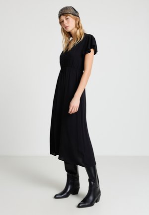 Donna che indossa un vestito midi nero con maniche svolazzanti, stivali neri al polpaccio e un foulard nero con motivo, in piedi davanti a uno sfondo semplice.