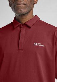 Bordeauxpoloshirt van een gestructureerde stof, met een kraag en een knoopsluiting met twee knopen, en een wit geborduurd logo op de linkerkant van de borst.